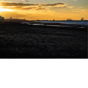 Warme zonsondergang boven een strand met silhouetten van gebouwen en havenkranen op de achtergrond.