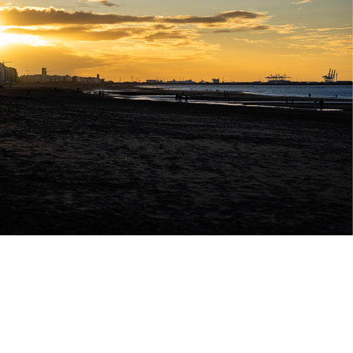 Warme zonsondergang boven een strand met silhouetten van gebouwen en havenkranen op de achtergrond.