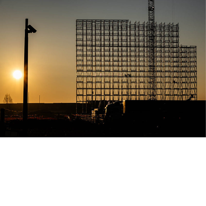 Foto van een metalen constructie in silhouet tegen een warme zonsopgang, met zon laag aan de horizon.
