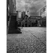 Zwart-witfoto van een fabriek met oude bakstenen gebouwen en moderne silo’s, onder een dreigende lucht