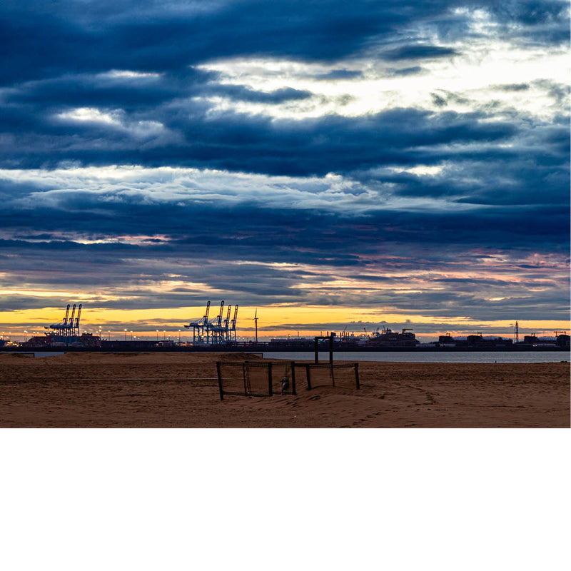 Foto van een strand met dramatische wolkenlucht en havenkranen op de achtergrond bij zonsondergang