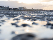 Foto van een strand met nat zand en reflecties van zonlicht, gebouwen op de achtergrond en zachte wolkenlucht.