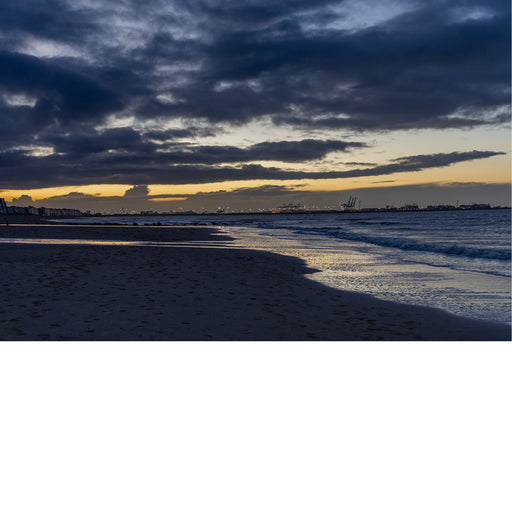 Foto van een strand bij schemering met dramatische wolken en lichtreflecties op het water.