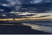 Foto van een strand bij schemering met dramatische wolken en lichtreflecties op het water.