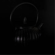 De foto toont een donkere gietijzeren theepot die subtiel uit een volledig zwarte achtergrond naar voren komt. 