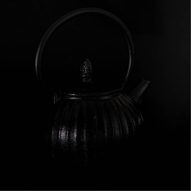 De foto toont een donkere gietijzeren theepot die subtiel uit een volledig zwarte achtergrond naar voren komt. 