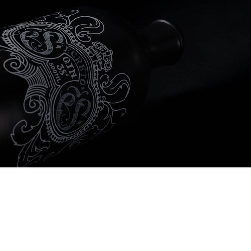 De foto toont een donkere, stijlvolle low-key opname van een fles gin, waarbij vooral het sierlijke etiket op de voorgrond komt. Door de minimale belichting worden enkel de fijne witte lijnen van het decoratieve ontwerp zichtbaar, terwijl de rest van de matte, zwarte fles bijna volledig in de schaduw verdwijnt.