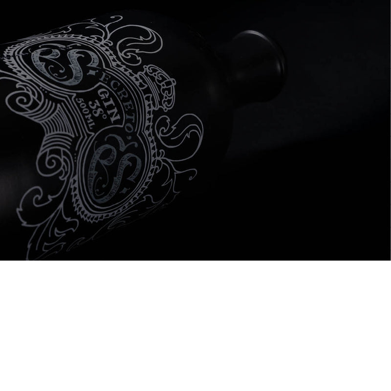 De foto toont een donkere, stijlvolle low-key opname van een fles gin, waarbij vooral het sierlijke etiket op de voorgrond komt. Door de minimale belichting worden enkel de fijne witte lijnen van het decoratieve ontwerp zichtbaar, terwijl de rest van de matte, zwarte fles bijna volledig in de schaduw verdwijnt.