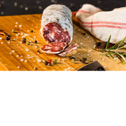 De foto toont een ambachtelijke droge worst die op een houten snijplank ligt, omringd door grove zoutkorrels, peper-ollen, kruiden en specerijen. 