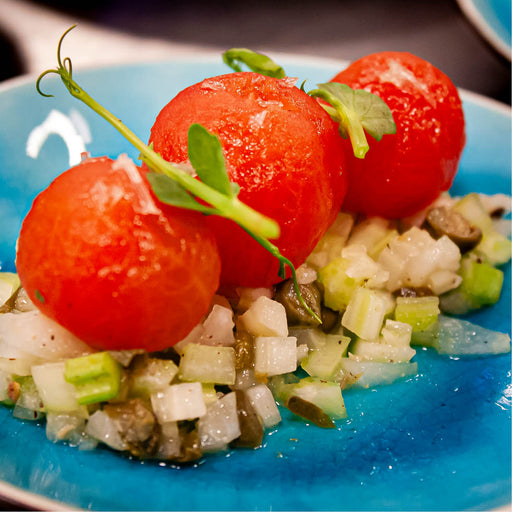 Fraai gestileerd culinair gerecht bestaande uit drie perfect gepelde, helderrode tomaatjes die elegant op een basis van fijngesneden groenten en kappertjes zijn geplaatst. 