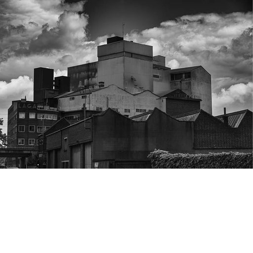 Massieve industriële gebouwen rijzen op tegen een dramatische wolkenlucht, waarbij de strakke metalen gevels sterk contrasteren met de oudere bakstenen structuren die op de voorgrond staan. 