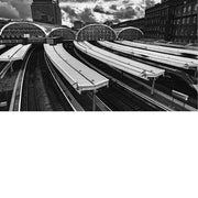 Onder een dramatische, zwaar gelaagde wolkenlucht ontvouwt zich een indrukwekkend spoorweglandschap waarin ritme en structuur de hoofdrol spelen. De gebogen stationsdaken met hun sierlijke metalen patronen vormen een opvallend contrast met de strakke, repetitieve lijnen van de perrons en rails. 