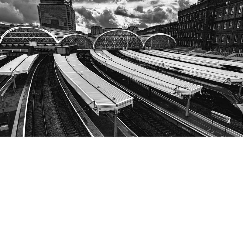 Onder een dramatische, zwaar gelaagde wolkenlucht ontvouwt zich een indrukwekkend spoorweglandschap waarin ritme en structuur de hoofdrol spelen. De gebogen stationsdaken met hun sierlijke metalen patronen vormen een opvallend contrast met de strakke, repetitieve lijnen van de perrons en rails. 