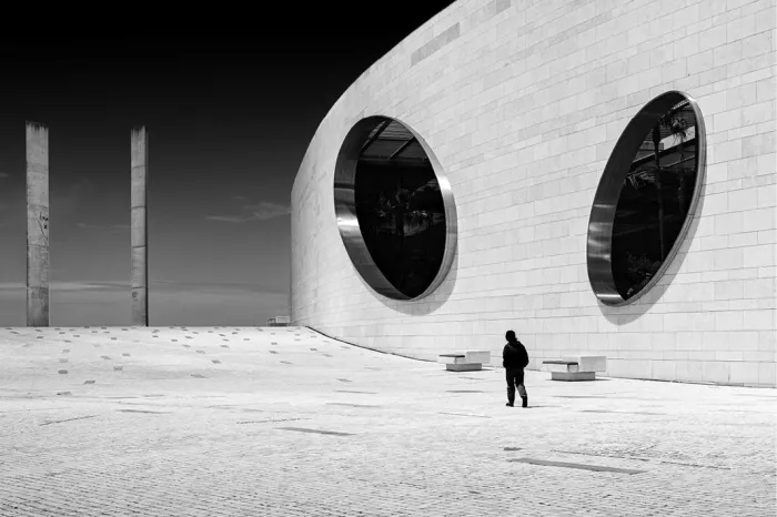 Wit gebogen gebouw van de Champalimaud Foundation in Lissabon met twee grote ronde vensters en een eenzame wandelaar op een zonovergoten plein, tegen een diepzwarte lucht.