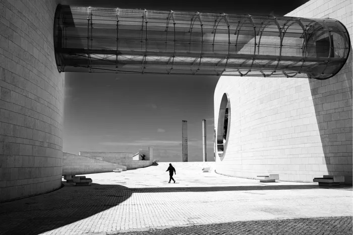 Glazen buisvormige loopbrug tussen de witte gebogen gevels van de Champalimaud Foundation in Lissabon, met een voorbijganger in het felle zonlicht en diepe slagschaduwen.