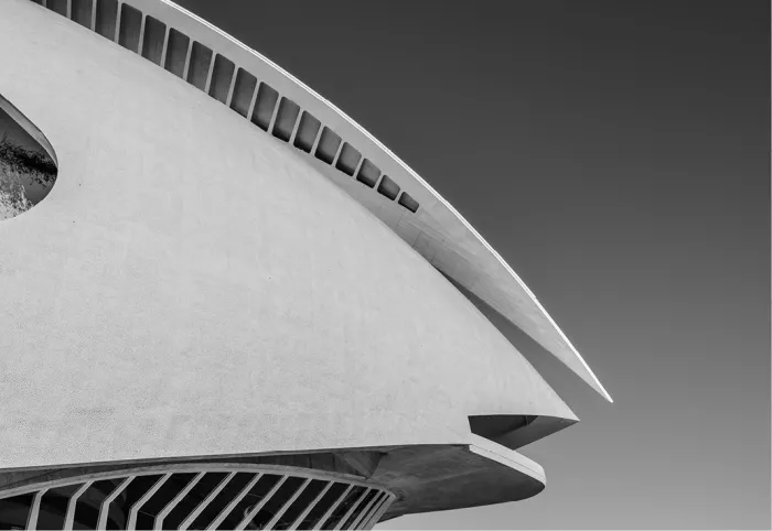 Wit, gebogen dak in de Ciutat de les Arts i les Ciències in Valencia steekt als een scherp blad uit boven een geribbelde gevel, tegen een egaal grijze lucht.