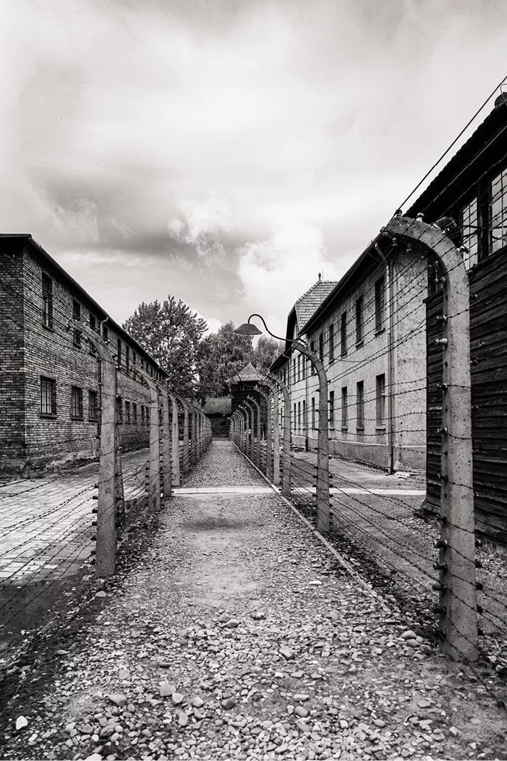 Grindpad tussen prikkeldraadomheiningen en bakstenen barakken in Auschwitz, met een wachttoren in de verte