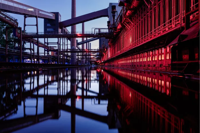 Cokesfabriek van Zollverein in Essen met rood verlichte gevel en stalen transportbanden, weerspiegeld in een waterbekken tijdens het blauwe uur.