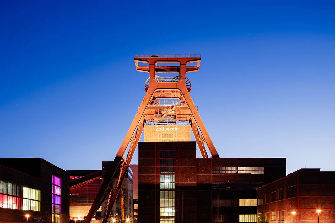 Oranje verlichte schachtbok van Zollverein boven bakstenen gebouwen tegen een diepblauwe avondlucht