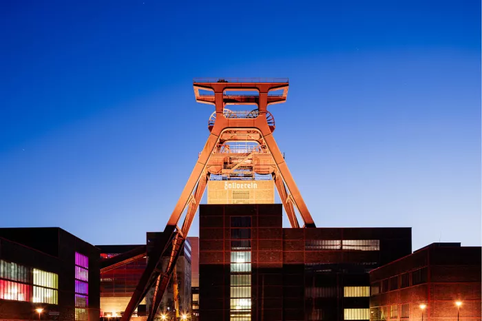 Oranje verlichte schachtbok van Zollverein boven bakstenen gebouwen tegen een diepblauwe avondlucht