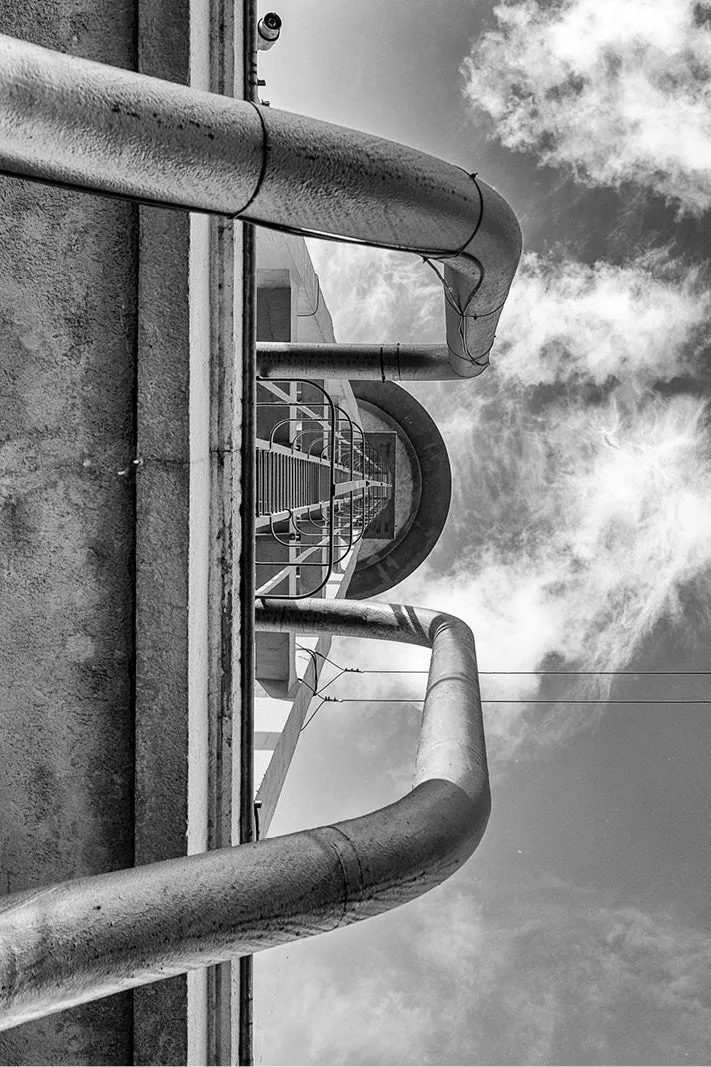 Gebogen metalen leidingen langs een betonnen toren, van onderen gezien tegen een lucht met witte wolken