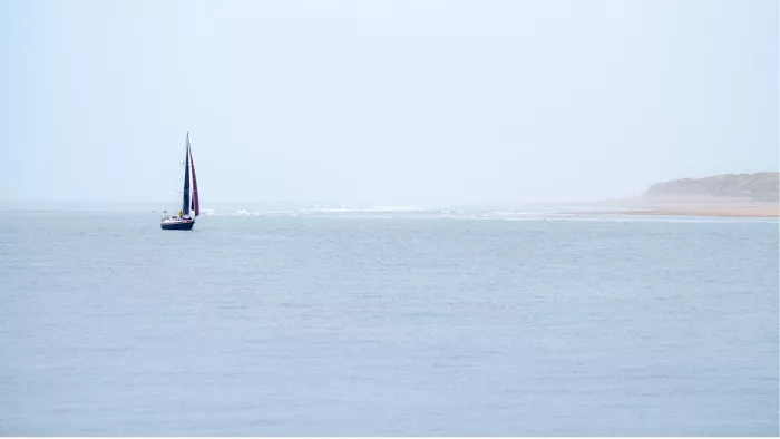 Kleine zeilboot met donkere zeilen op een kalme, lichtblauwe zee met duinen in de verte