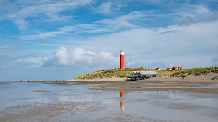 Rode vuurtoren op de duinen achter een breed strand, gespiegeld in een ondiepe strandplas