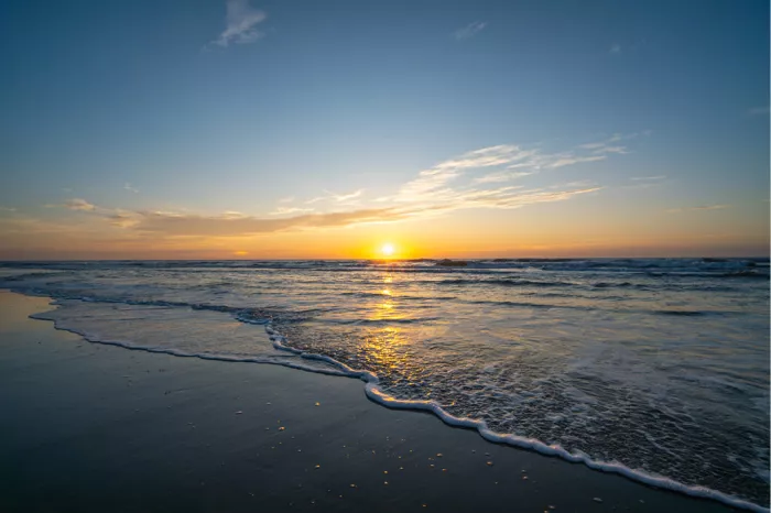 Een weids uitzicht over de zee tijdens zonsondergang. De zachte golven rollen over het natte zand en reflecteren de gouden lucht.