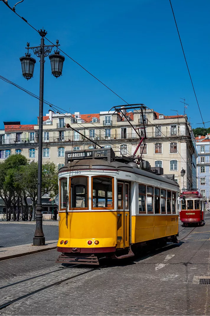 Een iconisch beeld van een felgele tram in een Europese stad, rijdend door een straat met oude, karaktervolle gebouwen onder een strakblauwe lucht.