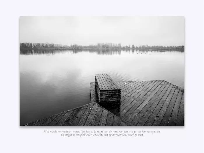 Een melancholisch landschap in zwart-wit. Een houten steiger strekt zich uit in kalm, spiegelglad water, omhuld door lichte mist op de achtergrond.