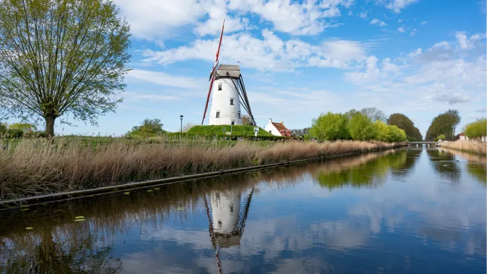 Een klassiek Hollands of Vlaams polderlandschap waarbij een grote witte windmolen perfect en stil wordt weerspiegeld in het water van een kaarsrecht kanaal.