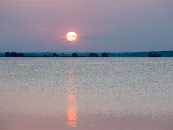 Gloeiende zon hangt laag boven een rustig wateroppervlak en werpt een lange rode weerspiegeling over het water, met een donkere bomenrand aan de horizon.