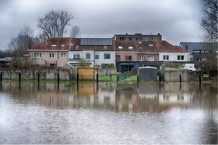 Achtertuinen en huizen met rode daken weerspiegelen in een grote regenplas in Merchtem onder een grijze lucht