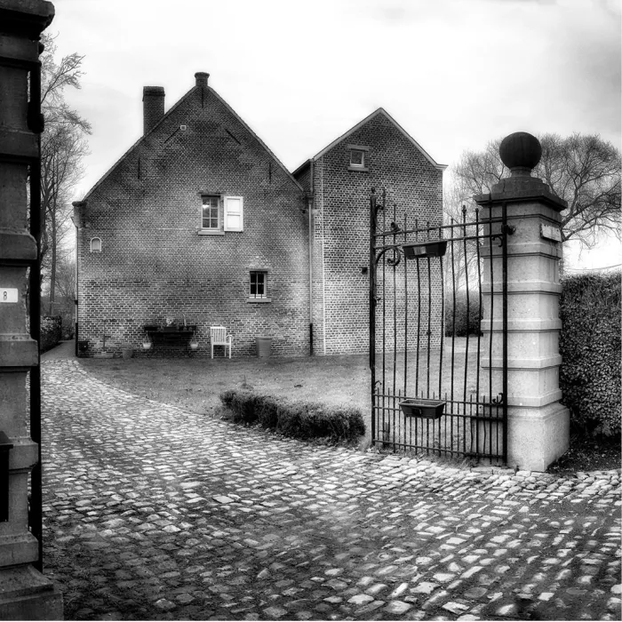 Zwart-wit foto van een bakstenen huis met kasseioprit, stenen poortpijlers en een open smeedijzeren poort