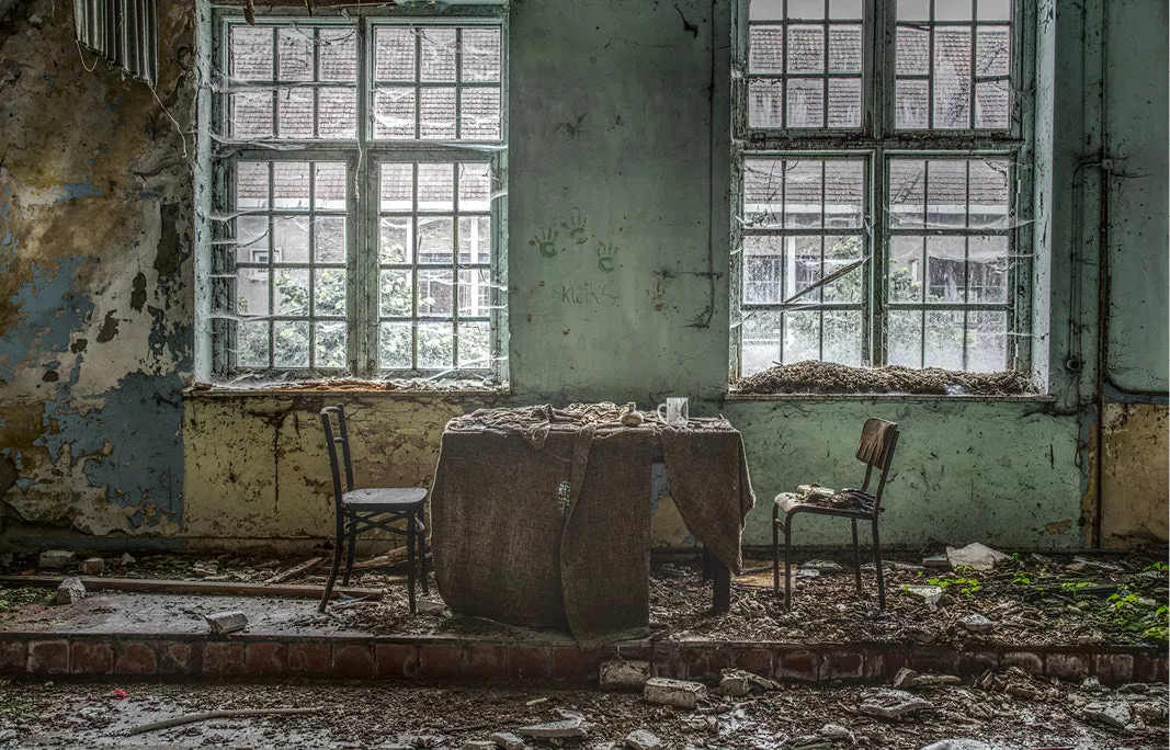 Vervallen theaterruimte met een tafel onder een doek, twee stoelen, afbladderende groene muren en puin onder twee grote ramen