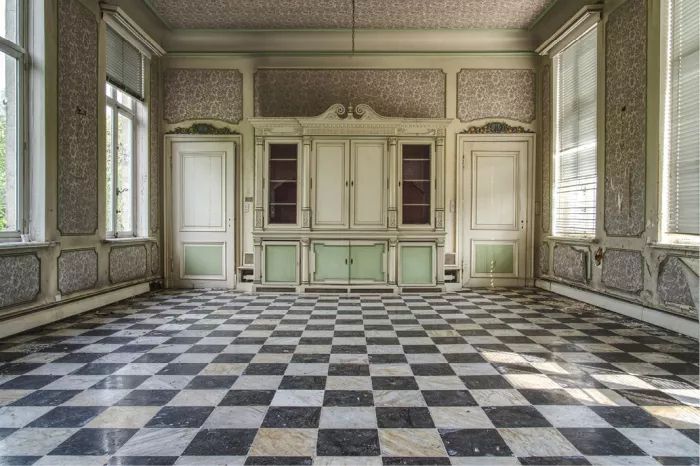 Lege woonkamer in een verlaten herenhuis met dambordvloer, een witte kast met groene panelen en daglicht door hoge ramen