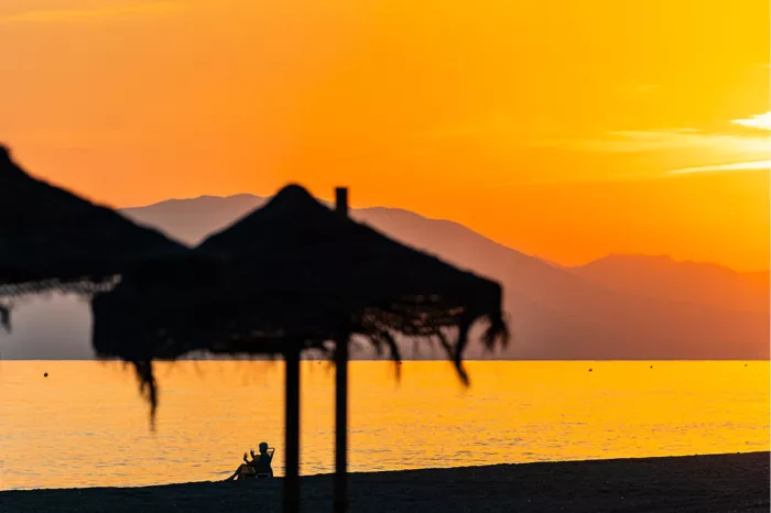 Silhouetten van strooien parasols en een zittende figuur op het strand bij een oranje zonsondergang met bergen
