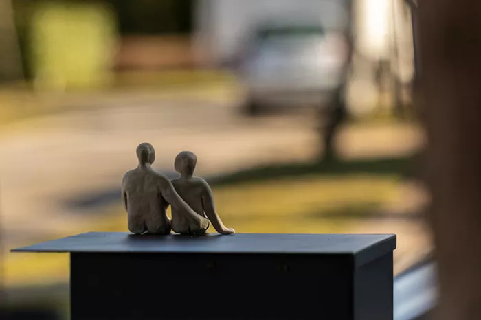 Sculptuurtje van twee zittende figuren naast elkaar op een sokkel, met een wazige straat erachter
