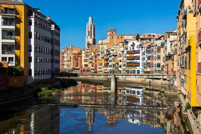 Kleurrijke gevels langs de rivier in Girona met een kerktoren en een brug, gespiegeld in het water