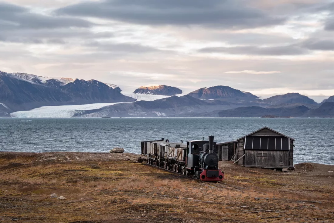 Zwarte stoomlocomotief met open wagens naast een houten schuur aan een fjord, met gletsjer en bergen erachter.