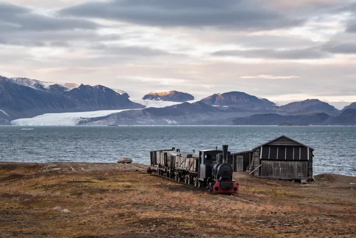 Zwarte stoomlocomotief met open wagens naast een houten schuur aan een fjord, met gletsjer en bergen erachter.