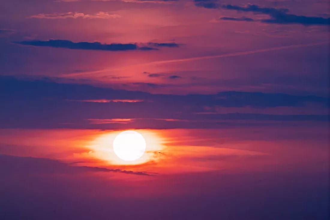 Felwitte zon met oranje gloed tussen banden van roze en paarse wolken bij zonsondergang.