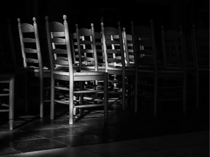 Lange rijen lege houten stoelen in een donkere kerk, met strijklicht over de ruggen en de vloertegels