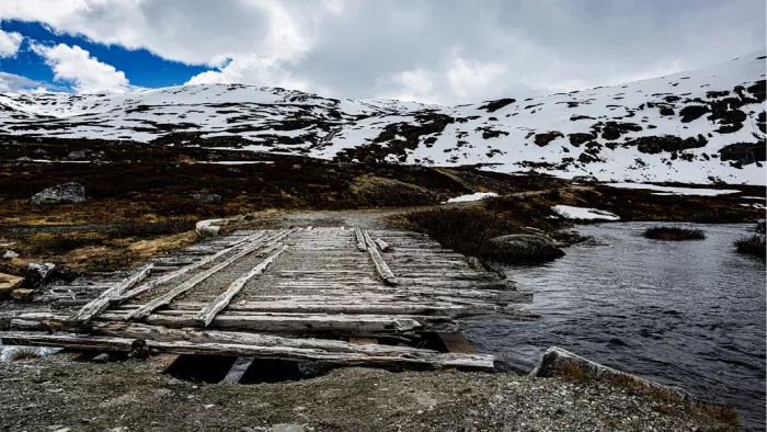 Vervallen houten brugje over donker water met besneeuwde heuvels erachter