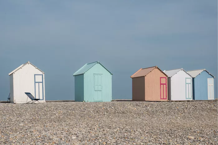 Rij kleurrijke strandhuisjes met puntdaken staat op een kiezelstrand aan de Baai van de Somme, onder een effen blauwe lucht.