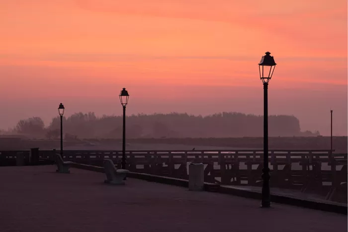 Straatlantaarns en bankjes staan op een lege promenade aan de Baai van de Somme, tegen een roze-oranje avondlucht met opkomende mist.