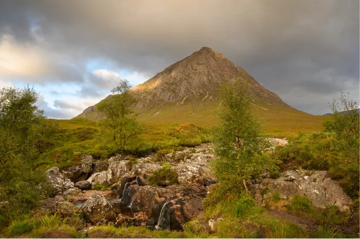 Kleine watervalletjes stromen tussen rotsblokken op de voorgrond, met de karakteristieke piek van Buachaille Etive Mor op de achtergrond onder een bewolkte lucht.