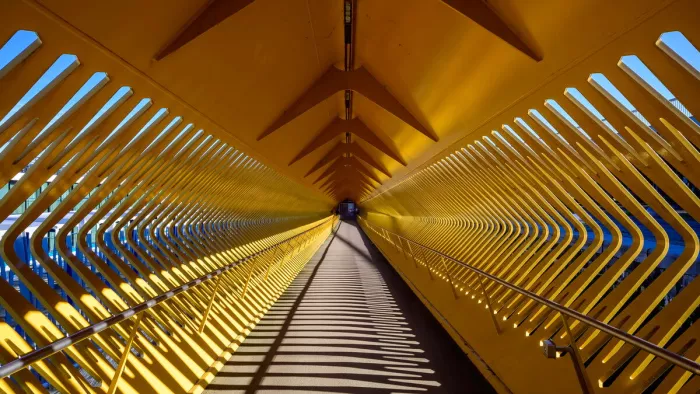 Gele loopbrug in Antwerpen met schuine spanten en strepen schaduw op de vloer die naar het einde lopen.