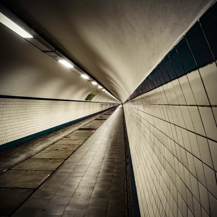 Lange voetgangerstunnel met witte tegelwanden en een rij lampen aan het gewelfde plafond die naar een verre uitgang toelopen.