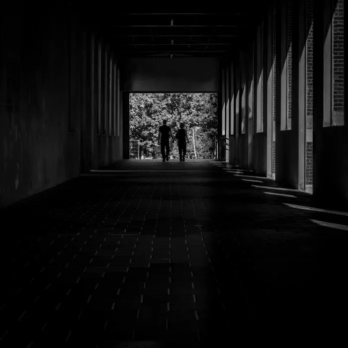 Twee mensen hand in hand in een donkere gang met pilaren, als silhouet tegen een lichte opening met bomen.